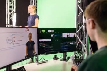  Blick in eine laufende Videoaufzeichnung. Eine Person im Hintergrund steht vor einem Greenscreen, eine andere Person im Vordergrund kontrolliert die Aufnahmen am Bildschirm 